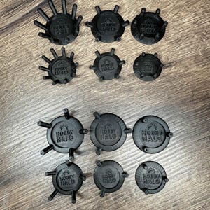 May include: A selection of black Hobby Halo accessories, circular in shape, with a central logo and radiating arms. The items are displayed on a wooden surface, highlighting their design and texture.