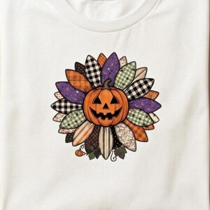 Peut inclure: Un t-shirt blanc avec un motif central représentant une citrouille jack-o'-lantern entourée de pétales de fleurs stylisés. Chaque pétale présente un motif différent, notamment du vichy noir et blanc, des rayures orange, du violet avec des lunes et du vichy vert.