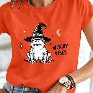 Peut inclure: Un t-shirt orange vif à manches courtes orné d'un motif noir et blanc représentant une grenouille portant un chapeau de sorcière. Le design inclut des étoiles, un croissant de lune et le texte noir "WITCHY VIBES" imprimé à droite de la grenouille.