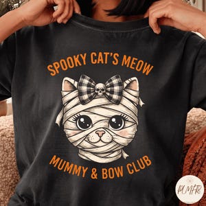 Peut inclure: Un t-shirt noir avec une illustration de chat enveloppé dans des bandages de momie, portant un nœud à carreaux noir et blanc avec un petit crâne. Le texte SPOOKY CAT'S MEOW et MUMMY & BOW CLUB est écrit en orange au-dessus et en dessous.
