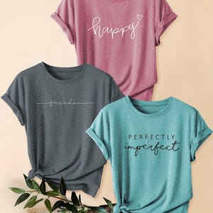 Peut inclure: Trois t-shirts à manches courtes en mauve, gris anthracite et bleu sarcelle. Le t-shirt mauve porte l'inscription "happy" avec un cœur. Le t-shirt gris affiche "freedom" en écriture cursive. Le t-shirt bleu sarcelle porte "PERFECTLY imperfect".