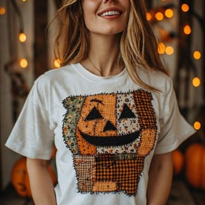 Peut inclure: Un t-shirt blanc à manches courtes arborant un grand motif central représentant une citrouille d'Halloween composée de divers tissus en patchwork aux tons orange, marron et vert, avec des contours noirs cousus formant le visage.