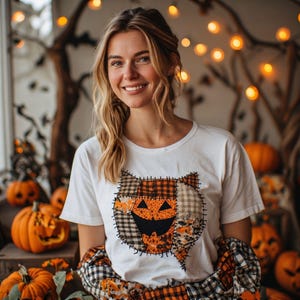 Peut inclure: Un t-shirt blanc à manches courtes avec un motif central représentant une tête de chat composée de divers empiècements en tissu à carreaux et à motifs orange, noirs et beiges, soulignés par des coutures noires visibles, porté avec une chemise en flanelle.