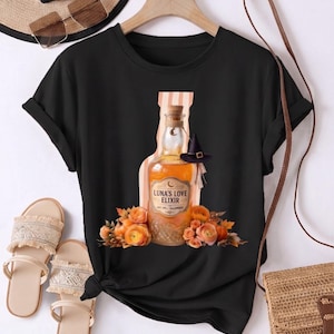 Peut inclure: Un t-shirt noir à manches courtes avec une impression graphique d'une bouteille en verre étiquetée Luna's Love Elixir. La bouteille est entourée de fleurs orange et de citrouilles, avec un petit chapeau de sorcière noir sur le goulot.