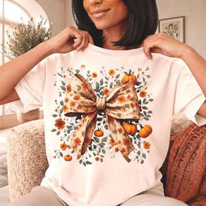 Peut inclure: Un t-shirt à manches courtes de couleur crème arborant un grand motif central représentant un nœud à imprimé floral. Le nœud est entouré d'une composition décorative de petites citrouilles orange et de feuillage vert, évoquant l'automne.