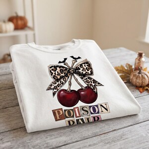 Peut inclure: Un sweat-shirt blanc à col rond orné d'un motif représentant deux cerises rouge foncé aux tiges en forme d'os, surmontées d'un grand nœud à imprimé léopard et de deux petites chauves-souris noires. Le texte POISON PAIR est imprimé en dessous.