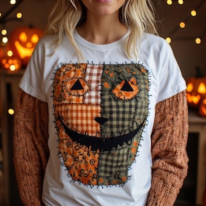 Peut inclure: Un t-shirt blanc à manches courtes arborant un grand motif central représentant un visage de citrouille en patchwork. Le design est composé de divers tissus orange et verts, incluant des motifs vichy et floraux, avec des détails cousus noirs.