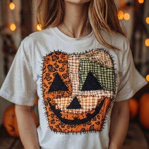 Peut inclure: Un t-shirt blanc à manches courtes arborant un grand motif central de citrouille composé de divers empiècements en tissu aux motifs orange, noirs et verts. Le design inclut des yeux triangulaires, un nez et une bouche noirs cousus.