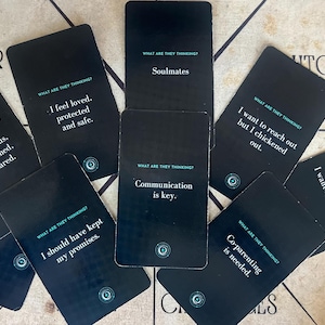 May include: A collection of black cards with white text, scattered on a light-colored surface. Each card features a phrase like "Soulmates" or "Communication is key." The cards are from a deck titled "What Are They Thinking?"