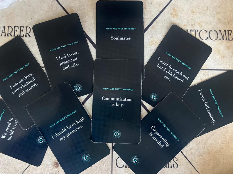 May include: A collection of black cards with white text, scattered on a light-colored surface. Each card features a phrase like "Soulmates" or "Communication is key." The cards are from a deck titled "What Are They Thinking?"