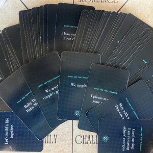 May include: A deck of black cards with white and teal text. The cards have rounded corners and feature phrases like "I love you" and "Let's build a life together." The deck is spread out on a light-colored surface.