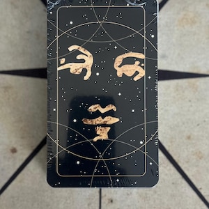 May include: A tarot card deck, sealed in plastic. The black card face has a gold face design, a starry background, and gold lines. The deck is rectangular with rounded corners.