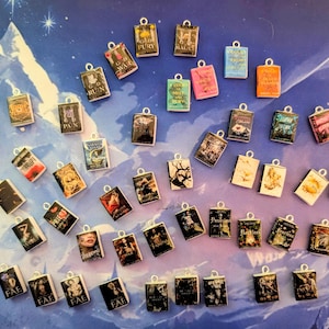Può includere: Una collezione di ciondoli a forma di libro in miniatura con anelli color argento per la creazione di gioielli. I ciondoli presentano varie copertine di libri con titoli come "Onyx Storm" e "Fury". Lo sfondo è una superficie blu e viola con un motivo a stelle.