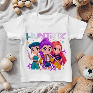May include: White short-sleeved t-shirt featuring a colorful graphic of three cartoon characters with the text "HUNTRIX". The characters have different hair colors and are holding stylized weapons. Stars and hearts surround the characters.