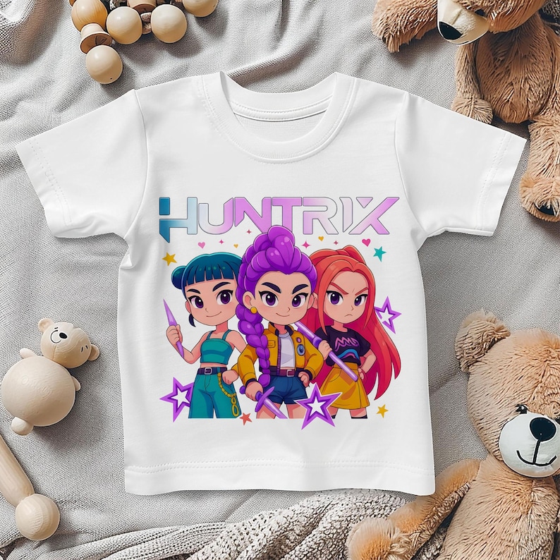 May include: White short-sleeved t-shirt featuring a colorful graphic of three cartoon characters with the text "HUNTRIX". The characters have different hair colors and are holding stylized weapons. Stars and hearts surround the characters.