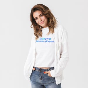 May include: A white t-shirt with the text "K-POP DEMON HUNTERS" in blue and purple. The person is wearing a white button-down shirt and blue jeans.