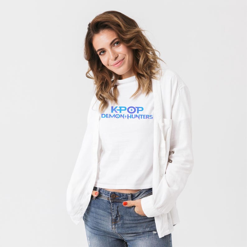 May include: A white t-shirt with the text "K-POP DEMON HUNTERS" in blue and purple. The person is wearing a white button-down shirt and blue jeans.