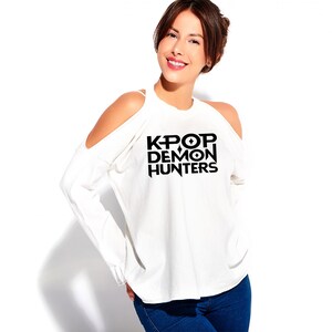 May include: White long-sleeve shirt with cold shoulder cutouts. The shirt features the black text "KPOP DEMON HUNTERS" in a bold, stylized font. The model is wearing blue jeans.
