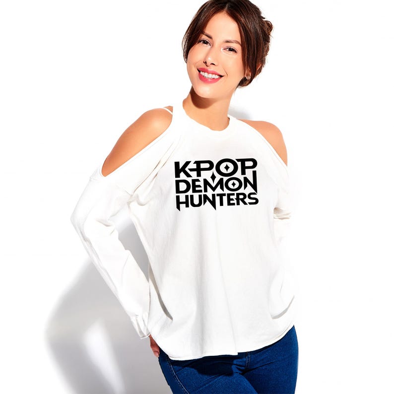 May include: White long-sleeve shirt with cold shoulder cutouts. The shirt features the black text "KPOP DEMON HUNTERS" in a bold, stylized font. The model is wearing blue jeans.