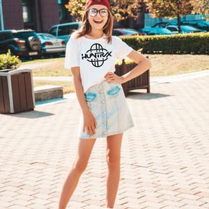 May include: A white t-shirt with the word "HUNTRIX" in black, paired with a light blue denim skirt. The outfit is completed with a red cap and glasses. The image is taken outdoors on a sunny day.
