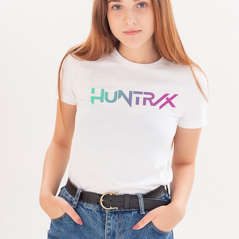 May include: White t-shirt with the word "HUNTR/X" in a gradient of teal, purple, and pink. The shirt has short sleeves and is paired with blue jeans and a black belt with a gold buckle.