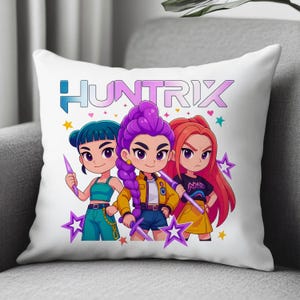 May include: White decorative pillow featuring a cartoon illustration of three female characters with the word "HUNTRIX" above them. The characters have different hair colors and hold stylized weapons, surrounded by stars.