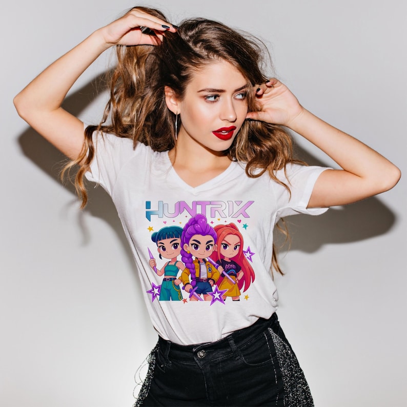 May include: White t-shirt featuring the word "HUNTRIX" in a colorful font above a graphic of three cartoon characters. The characters have different hair colors and are wearing various outfits. The shirt is a v-neck style.