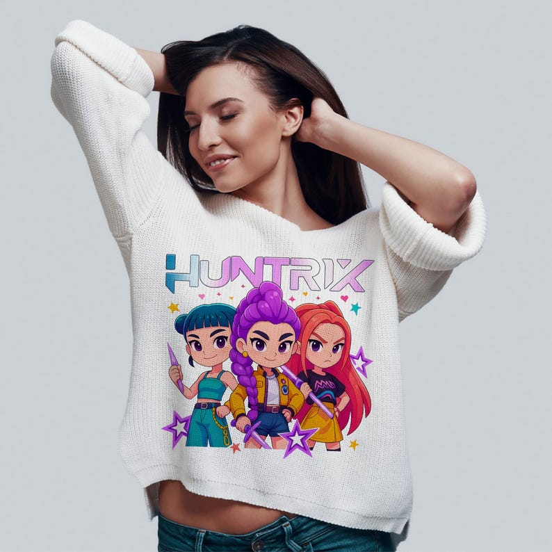 May include: White long-sleeve sweater featuring the word "HUNTRIX" in blue and pink. The graphic shows three cartoon characters with colorful hair and outfits, holding star-shaped wands. The sweater is a casual, everyday garment.