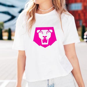 May include: White t-shirt featuring a vibrant pink lion graphic. The lion design is a stylized, geometric illustration with a bold, modern aesthetic. The shirt is a classic crew neck style.