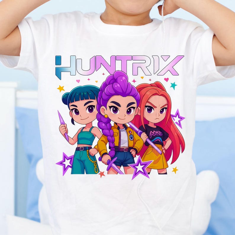 May include: White t-shirt featuring the word "HUNTRIX" in teal and a graphic of three cartoon girls with weapons. The girls have blue, purple, and red hair, and are surrounded by stars.