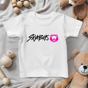 May include: White t-shirt with the text "SAJA BOYS" in black script and a pink lion logo. The shirt is made of a soft material and is suitable for children. The shirt is displayed on a light gray surface.