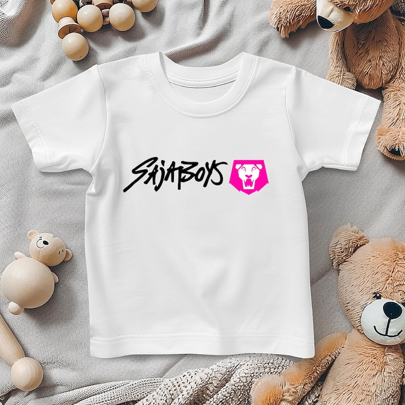 May include: White t-shirt with the text "SAJA BOYS" in black script and a pink lion logo. The shirt is made of a soft material and is suitable for children. The shirt is displayed on a light gray surface.