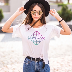 May include: White t-shirt with a colorful graphic that says "HUNTRAX". The graphic is a combination of pink, purple, and teal colors. The shirt is paired with blue jeans and a black belt.