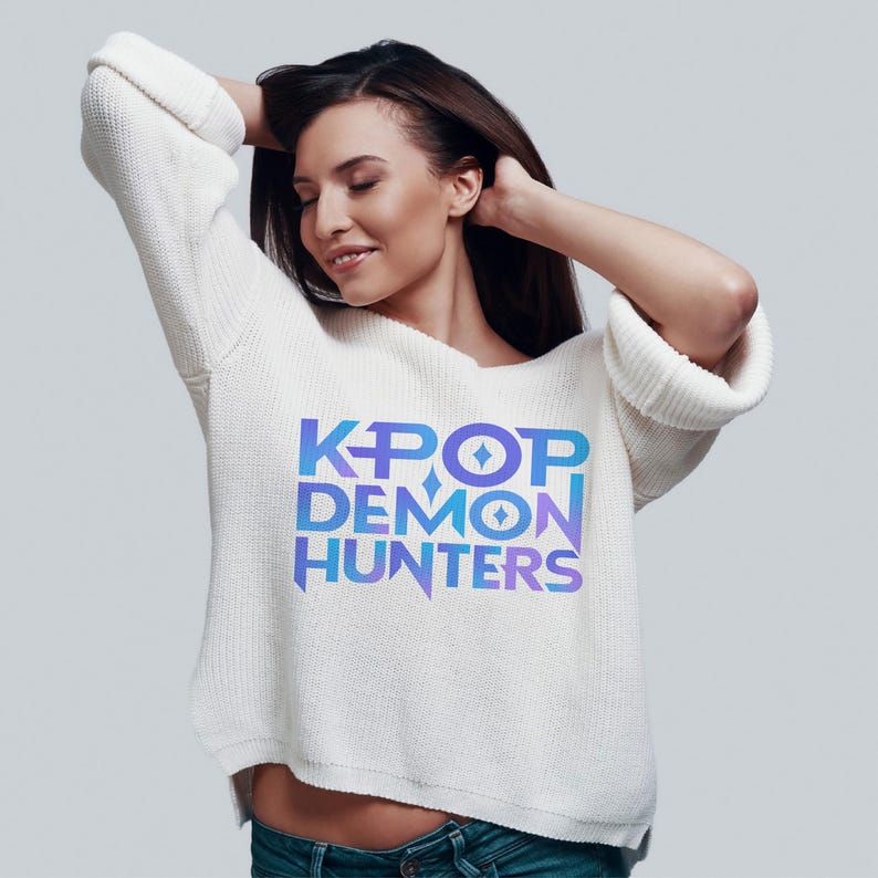 May include: A white sweater with the text "KPOP DEMON HUNTERS" in blue and purple gradient lettering. The sweater has long sleeves and a relaxed fit. The person in the image is wearing blue jeans.