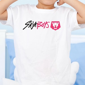May include: White t-shirt featuring the text "Saja Boys" in black and pink, with a lion logo in a pink shield. The shirt is a crew neck and appears to be made of cotton.