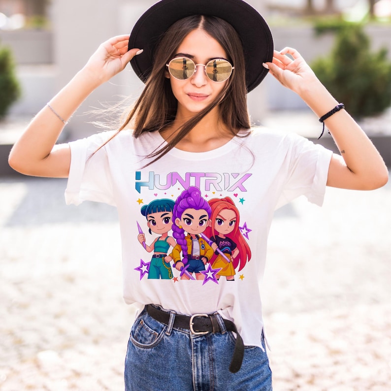 May include: White t-shirt featuring the word "HUNTRIX" in blue and purple, with a graphic of three cartoon characters. The characters have different hair colors and hold weapons. The shirt is worn with blue jeans and a black belt.