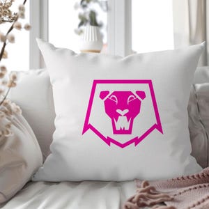 May include: White square throw pillow with a bright pink lion head design. The lion graphic is enclosed in a geometric frame. The pillow is on a bed with other white pillows and a pink blanket.
