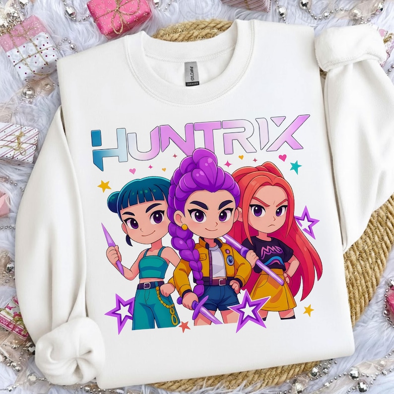 May include: White sweatshirt featuring the word "HUNTRIX" in a colorful font above a graphic of three cartoon characters. The characters have different hair colors and hold weapons. The design includes stars and hearts.