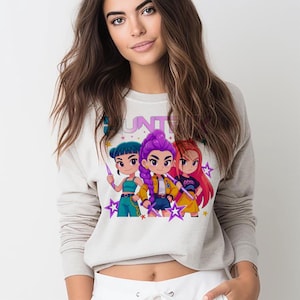 May include: A light gray sweatshirt featuring a colorful graphic of three anime-style characters holding glowing rods, with star accents. The word "HUNTER" is printed across the top. The characters are wearing different outfits and have unique hairstyles.