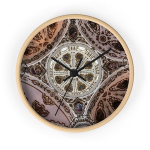 May include: A round wall clock with a light wood frame. The clock face features a detailed image of an ornate ceiling with gold and white architectural details. Black clock hands indicate the time.