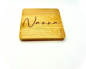 May include: A square bamboo coaster with the name "Nanna" engraved in a cursive font. The coaster is a light brown color and has visible wood grain.