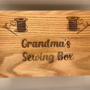 May include: A wooden sewing box lid with the words "Grandma's Sewing Box" engraved. The lid also features two decorative spools of thread and needles. The box is made of light-coloured wood.