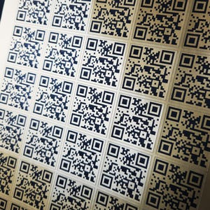 May include: Close-up of a sheet of gold-colored stickers printed with multiple black QR codes. The codes are arranged in a grid pattern, each a unique square of black and white pixels.
