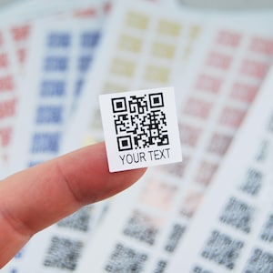 May include: A small, square white sticker with a black QR code and the text "YOUR TEXT" is held between fingers. Other stickers with QR codes in various colors are in the background.