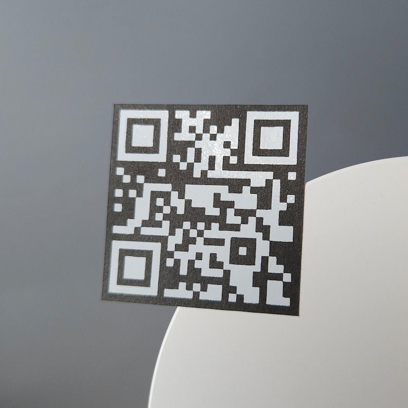 May include: A square QR code with a black and white pattern. The code is positioned on a white surface against a gray background. The code is composed of small black and white squares arranged in a unique pattern.