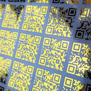 May include: Close-up of a sheet of stickers with gold QR codes on a light blue background. The QR codes are arranged in a grid pattern, with a shiny, metallic finish. The stickers are likely for product labeling or marketing.
