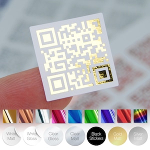 May include: A small, square white sticker featuring a gold QR code design. The sticker is held between fingers, with a selection of colour options below, including white matte, white gloss, clear gloss, clear matte, black stickers, gold matte, and silver matte.