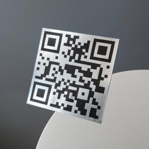 May include: A square, silver QR code with a black pattern. The code is displayed at an angle, with a white and gray background. The QR code is a modern design element.