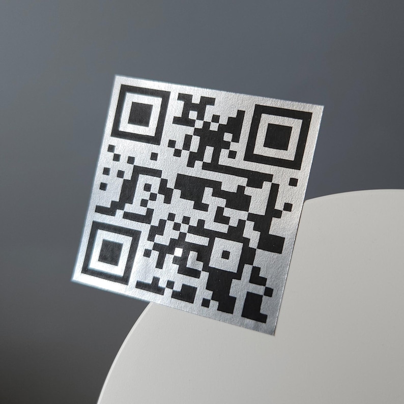 May include: A square, silver QR code with a black pattern. The code is displayed at an angle, with a white and gray background. The QR code is a modern design element.