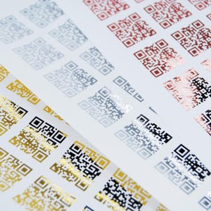 May include: Sheets of QR code stickers in various metallic colors, including gold, silver, and copper, arranged diagonally. The codes are printed on a white background, creating a contrast between the shiny codes and the matte paper.